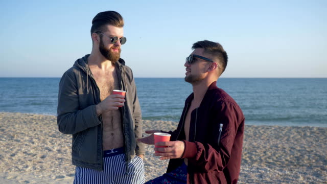 male gossip, cheerful friends in glasses are drinking tea and talking on beach