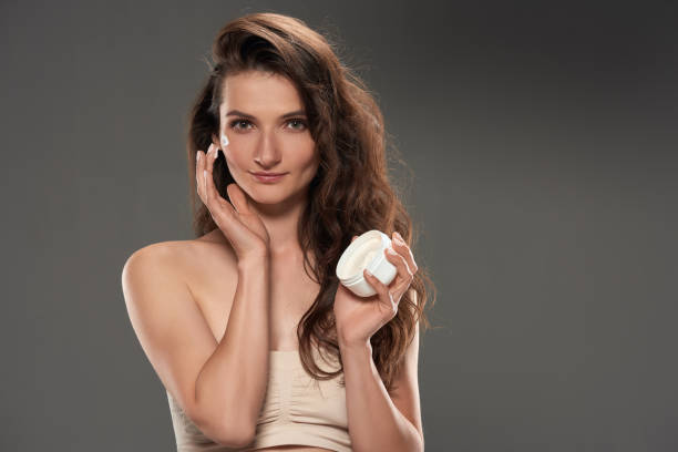 istock beautiful brunette woman applying moisturizing cream on face, isolated on grey 996220652