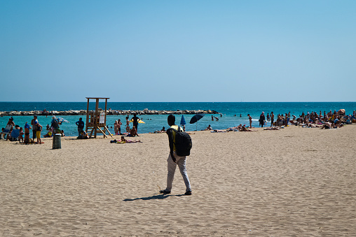 Bari,Italy,07/06/2018; one black boy with his shoulder bag a sunny morning on the beach of adriatic coast of Bari,who walks against the background of the crowd of bathers.