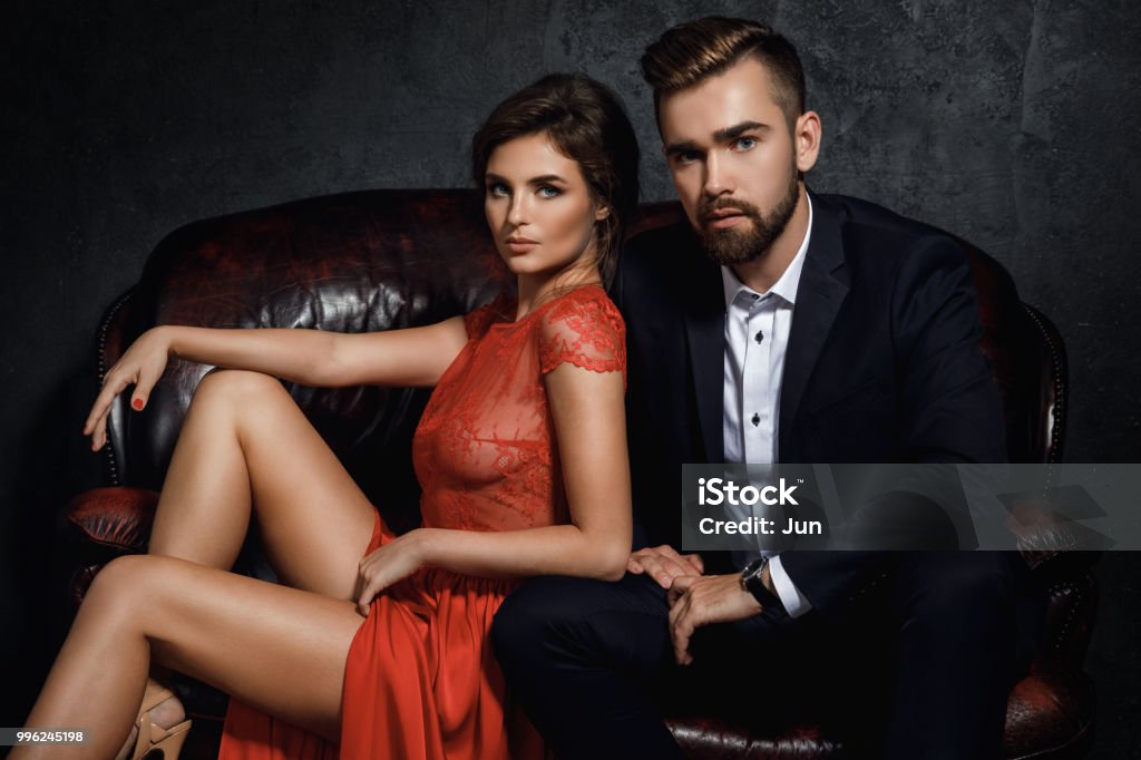Attractive young couple is sitting on the leather sofa Attractive young couple is sitting on the leather sofa. Woman in red dress and man wearing classical suit. Men Stock Photo