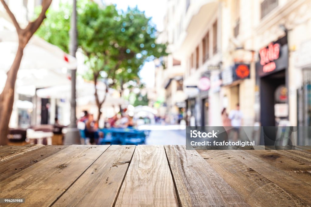 Empty wooden table with defocused sidewalk cafe at background Night view of an empty rustic wooden table in the foreground with defocused lights of a town street café at background ideal for product or food display. Predominant colors are brown and yellow. DSRL outdoors photo taken with Canon EOS 5D Mk II and Canon EF 24-105mm f/4L IS USM Wide Angle Zoom Lens Table Stock Photo