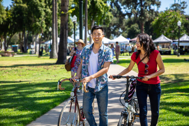 Los Angeles Couple Wheeling Bikes Through Park Durning Farmer's Market A man and woman are smiling and laughing as they walk through a park in Pasadena, CA. the stalls of a farmer's market can be seen in the background. They are pushing bicycles. farmers market healthy lifestyle choice people stock pictures, royalty-free photos & images