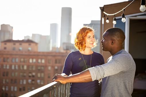 A man and woman in their 30s stand together on a balcony overlooking Downtown Los Angeles at sunset.