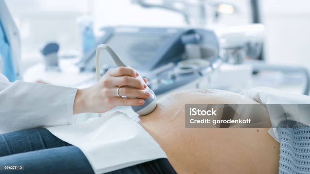 In the Hospital, Close-up Shot of the Doctor Doing Ultrasound / Sonogram Scan to a Pregnant Woman. Obstetrician Moving Transducer on the Belly of the Future Mother. Ultrasound Stock Photo