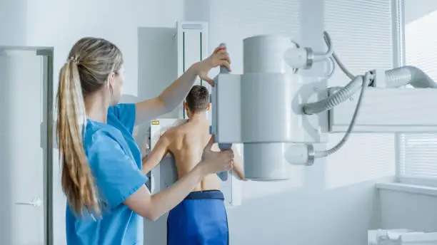 Photo of In the Hospital, Man Standing Face Against the Wall While Medical Technician Adjusts X-Ray Machine For Scanning. Scanning for Fractures, Broken Limbs, Chest, Cancer or Tumor. Modern Hospital with Technologically Advanced Medical Equipment.