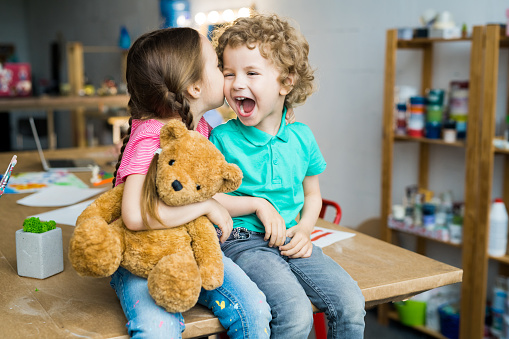 Warm toned portrait of two cute little kids kissing and laughing happily hugging teddy bear toy, copy space