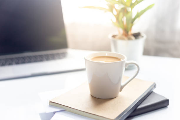 istock Coffee cup on notebook with laptop on white table in the morning sun light ,Working space 996341424