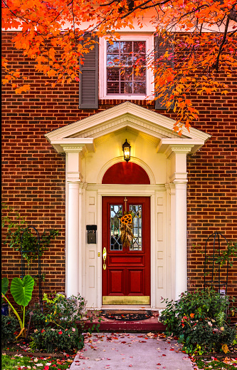 Entrance to brick house with pillars for porch and red door with autum leaves on sidewalk and colorful branches framing picture - fall flowers on leaded glass window in door