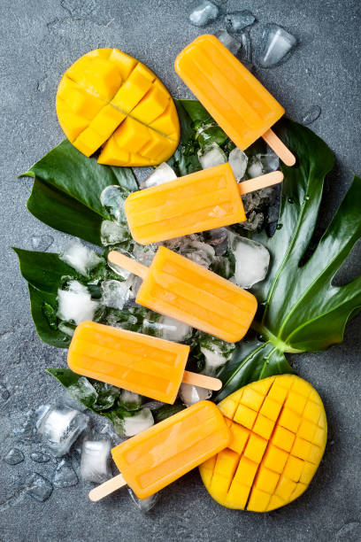 istock Mango popsicles over green tropical palm leaf on grey stone background. Overhead, top view 996472704