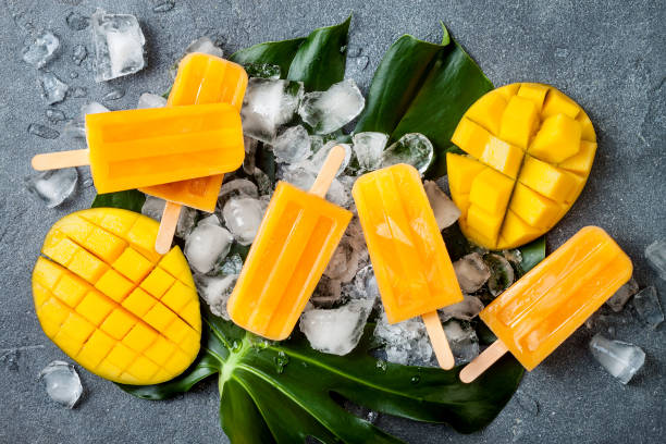 istock Mango popsicles over green tropical palm leaf on grey stone background. Overhead, top view 996472714