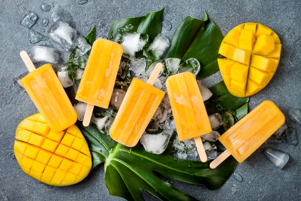 istock Mango popsicles over green tropical palm leaf on grey stone background. Overhead, top view 996472718