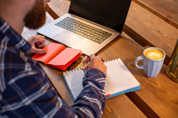 istock Cropped image of casual business man or freelancer planning his work on notebook, working on laptop computer with smart phone, cup of coffee on table at coffee shop or home office 996475506