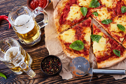 close-up of classic italian Pizza Margherita cut in slices on a parchments paper on a old rustic wooden table with beer in glass cups,  authentic recipe, view from above