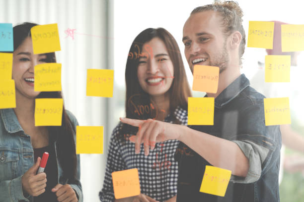 istock Group of young successful creative multiethnic team smile and brainstorm on project together in modern office with post note or sticky note. Caucasian man point on glass wall sharing idea. 996496574