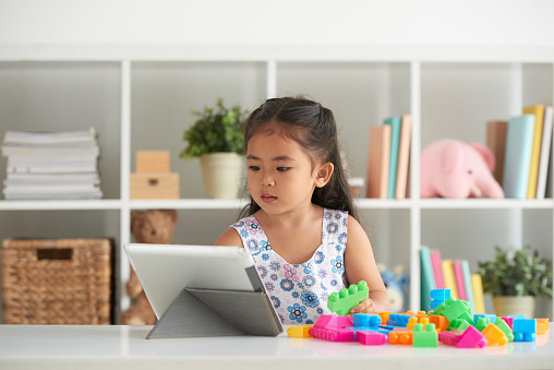 Little girl building a tower from blocks and using digital tablet at the table