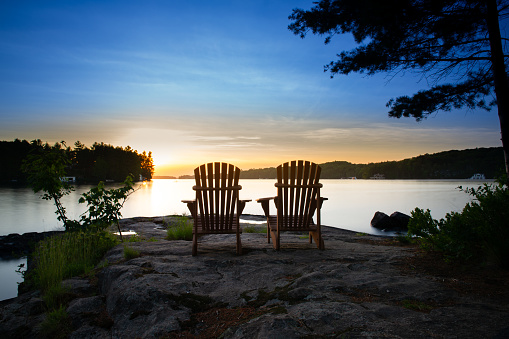 Two Muskoka chairs sitting on a rock formation facing a calm lake at sunset.