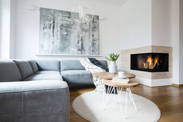 istock Grey corner settee with blankets standing in white Nordic living room interior with fresh tulips, books and tea cup on hairpin tables, abstract painting and fireplace 996692482