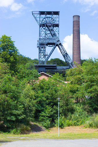 The mine tower for black coal mining Landek in city Ostrava in the Czech Republic. In the background is blue sky with white clouds. In foreground are trees and bushes and lamp.