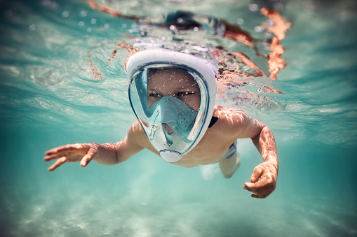 Happy little boy swimming underwater in the sea. The boy is wearing a modern full-face snorkel mask.
Nikon D850