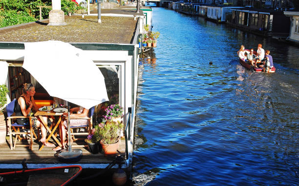 Two women having lunch on the terrace of a traditional houseboat on the canals during a sunny and warm summer day in Amsterdam.          A Canal cruise boat passing by makes the scene almost idyllic.\nHouseboats in Amsterdam are generally not motorized, but moored to a fixed spot in the city. The intricate network of canals in Amsterdam’s city centre also serves as a unique residential area. The colourful houseboats moored along canal banks throughout the city offer all the comforts of a typical house and have become something of a local icon. There are around 2,500 houseboats in Amsterdam.