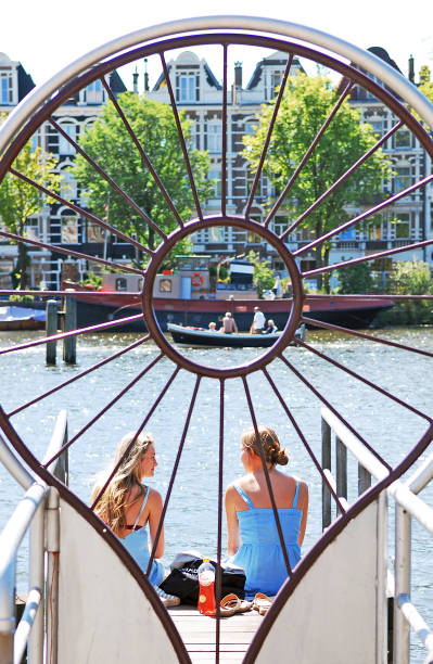 Two happy young women talking on the Amstel river pier. Two happy young women relax and talk while having a lunch sitting together on one of the many piers on the Amstel river. They are peacefully enjoying a breathtaking view of the canal houses of Amsterdam. jordaan amsterdam stock pictures, royalty-free photos & images