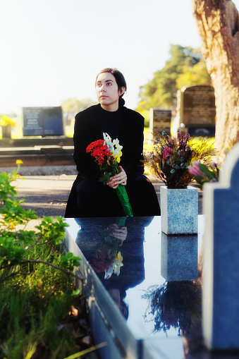 A bereaved young woman walks sadly through a cemetery carrying a bunch of flowers to the grave of a loved one.