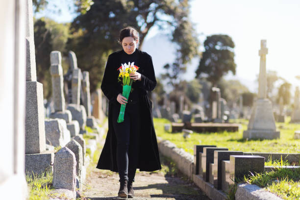 Bereaved young woman in black taking flowers to grave A young woman in black walks through cemetery headstones carrying flowers to the grave of someone she misses. funeral photos stock pictures, royalty-free photos & images