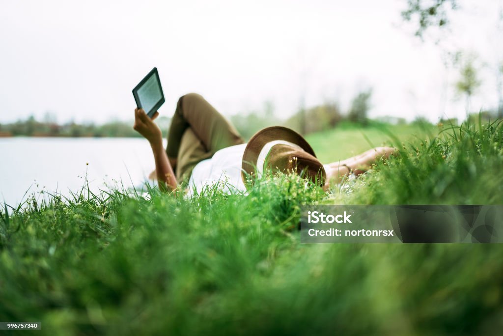 Godersi l'e-book vicino al lago. Ragazza sdraiata nell'erba. - Foto stock royalty-free di Leggere