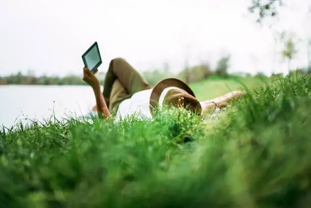 Enjoying e-book near the lake. Girl lying in the grass.