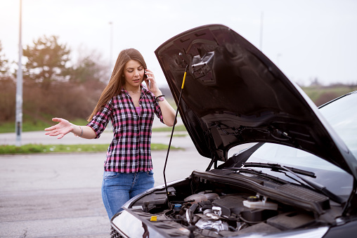 Young worried girl is using a phone to call the mechanic to fix the car whos hood she is looking under.