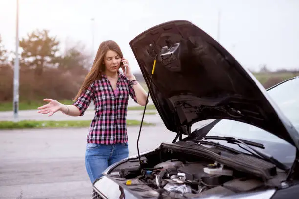 Photo of Young worried girl is using a phone to call the mechanic to fix the car whos hood she is looking under.