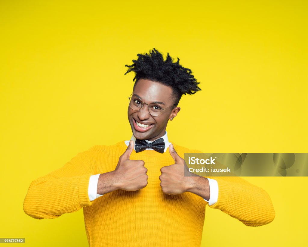 Funny portrait of nerdy young man with thumbs up Funny portrait of nerdy young afro American man wearing yellow sweater and black bow tie laughing at the camera with thumbs up. Studio Shot Stock Photo