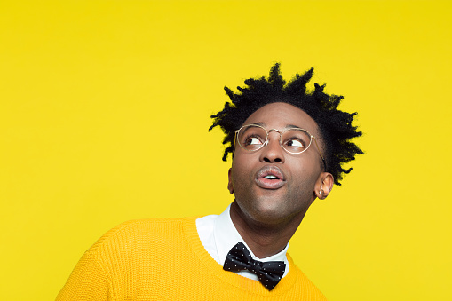 Funny portrait of surprised cheesy nerdy young afro American man wearing yellow sweater, black bow tie and glasses, looking up.