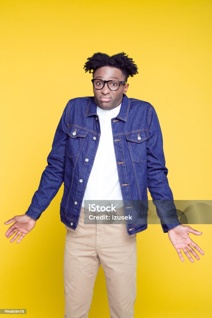 80's style portrait of confused nerdy young man Portrait of worried nerdy young afro American man wearing oversized jeans jacket and glasses, standing against yellow background. Confusion Stock Photo