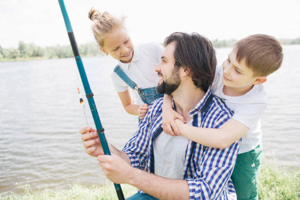 istock Kids are standing behind their dad, hugging and looking at him while man is holding fish-rod and looking at daughter. They are standing at river shore. They are happy 996811980