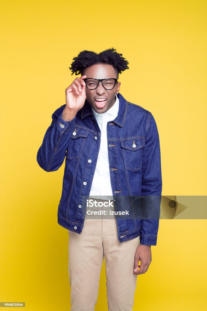 80's style funny portrait of nerdy young man Funny portrait of nerdy young afro american man wearing oversized jeans jacket and glasses, standing against yellow background. Bizarre Stock Photo