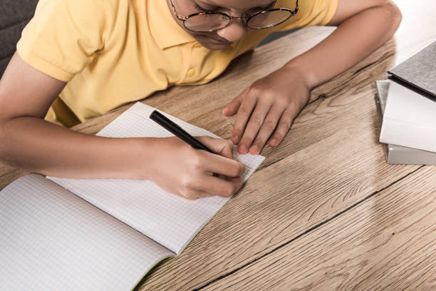 istock partial view of schoolboy in eyeglasses writing in empty textbook at table with stack of books 996813128
