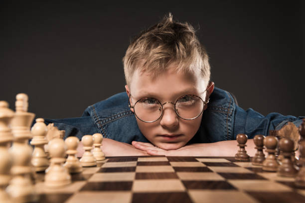 istock serious little boy in eyeglasses looking at camera and sitting at table with chess board isolated on grey background 996813320