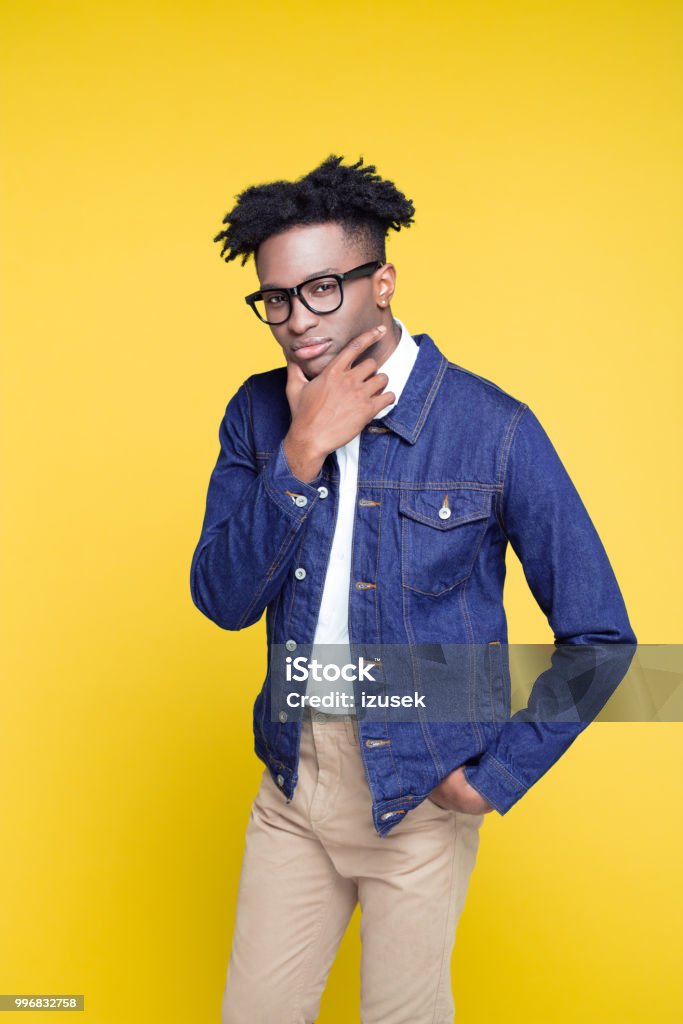 80's style portrait of confident nerdy young man Portrait of nerdy young afro American man wearing oversized jeans jacket and glasses, standing against yellow background. Contemplation Stock Photo