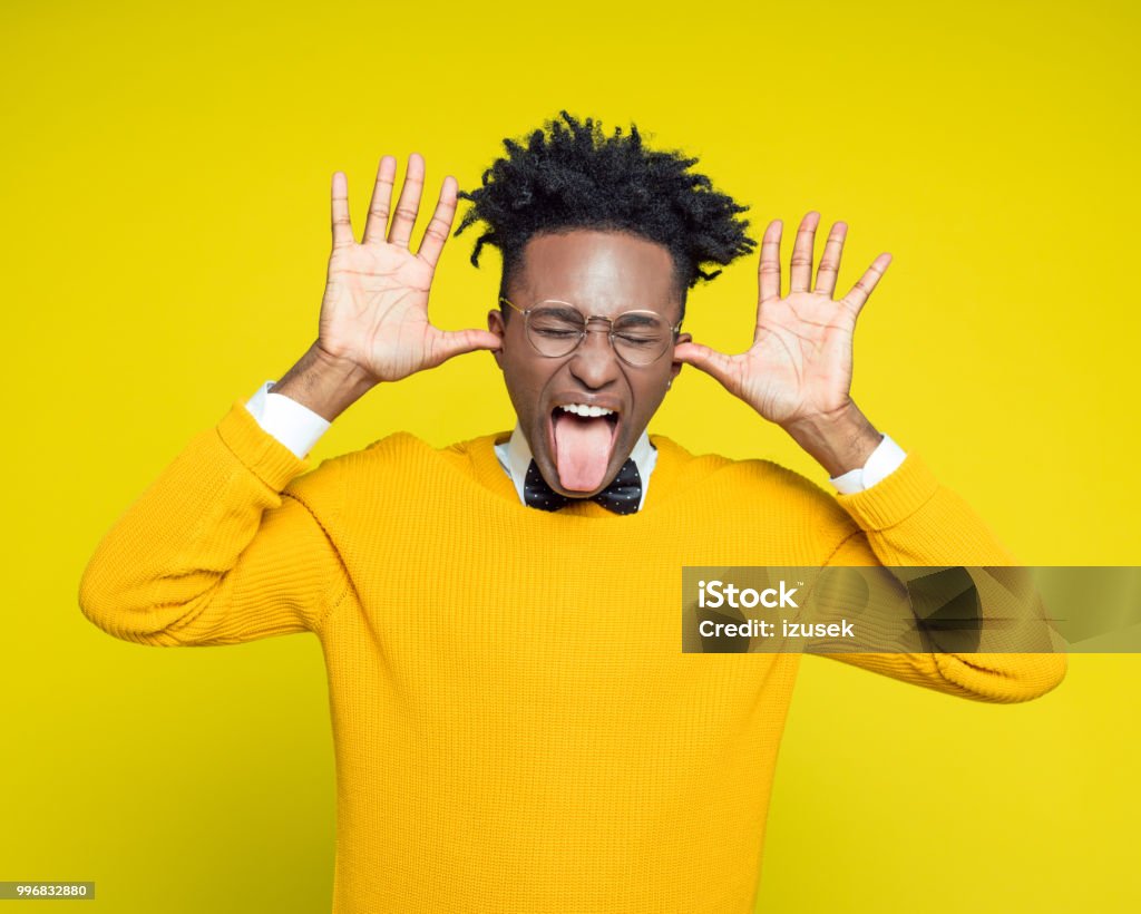 Funny portrait of nerdy young man sticking out tongue Funny portrait of nerdy young afro American man wearing yellow sweater and black bow tie sticking out tongue, standing against yellow background. 1980-1989 Stock Photo