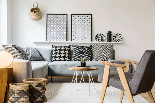 Photo of Real photo of a small table standing between a sofa with pillows and an armchair on a white rug in boho living room interior with posters on a shelf