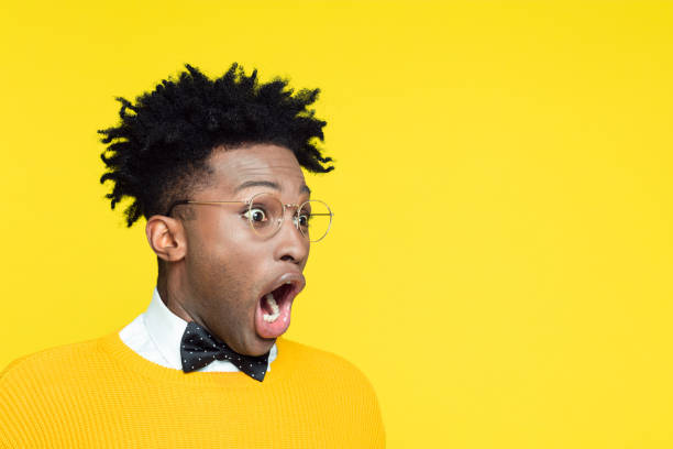 Portrait of shocked nerdy young afro american man wearing yellow sweater and black bow tie looking at copy space with mouth open against yellow background.