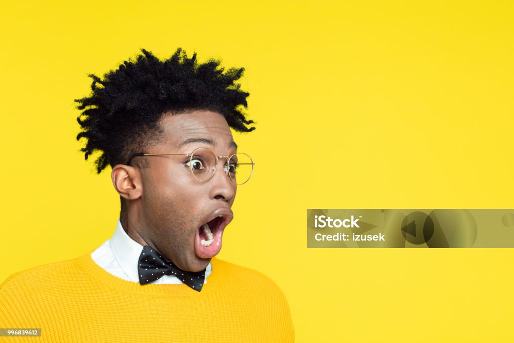 Portrait of surprised geeky young man looking away Portrait of shocked nerdy young afro american man wearing yellow sweater and black bow tie looking at copy space with mouth open against yellow background. Surprise Stock Photo
