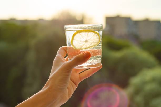 glass of clear, sparkling water with lemon in hand. background sky, silhouette of the city, sunset - sky human hand water white imagens e fotografias de stock