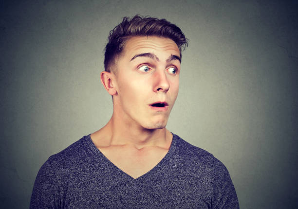 Young casual man looking super amazed while eavesdropping and looking away on gray background
