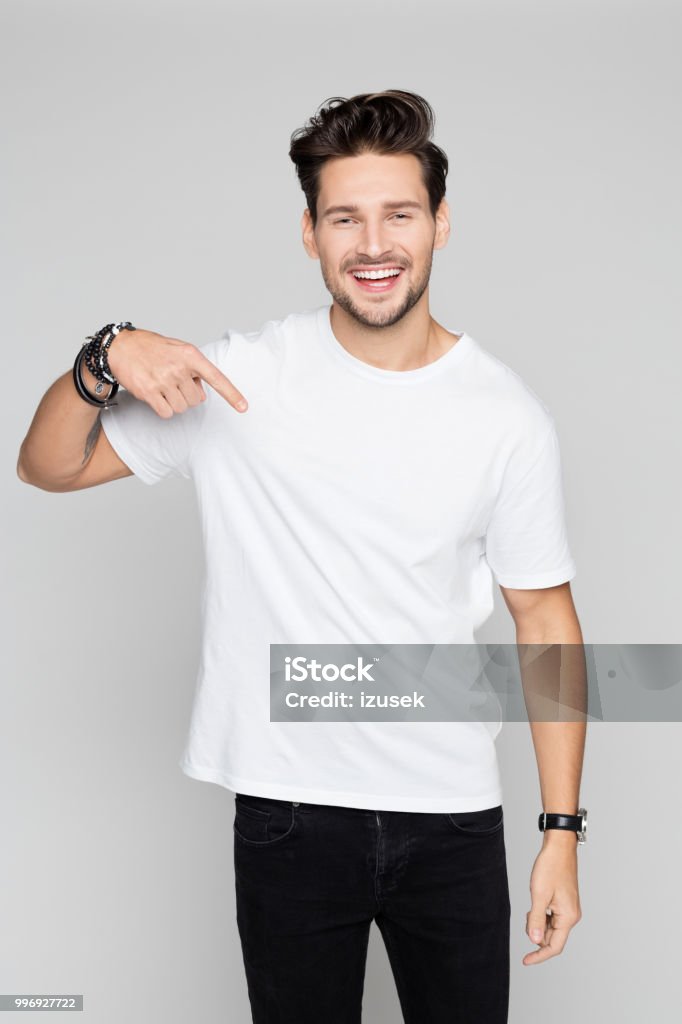 Smiling young man pointing down Portrait of young man pointing down and smiling on grey background T-Shirt Stock Photo