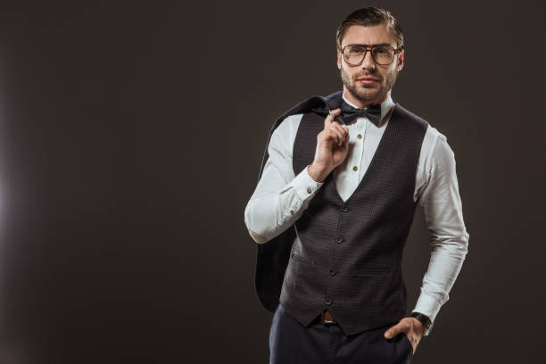 handsome stylish man in bow tie and eyeglasses holding suit jacket and looking at camera isolated on black