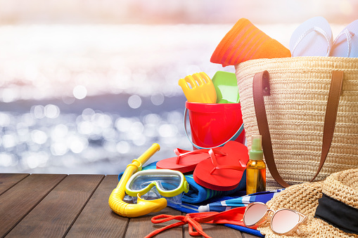 Front view of multi colored beach accessories shot on wooden plank with defocused beach at background. Accessories included in the composition are a beach umbrella, beach towels, a pair of flip-flops, scuba mask with snorkel, suntan lotion, beach bag, sun hat and a sunglasses. The accessories are grouped at the right of the frame leaving useful copy space for text and/or logo. Beach, summer or vacations concept. DSRL outdoors photo taken with Canon EOS 5D Mk II and Canon EF 24-105mm f/4L IS USM Wide Angle Zoom Lens