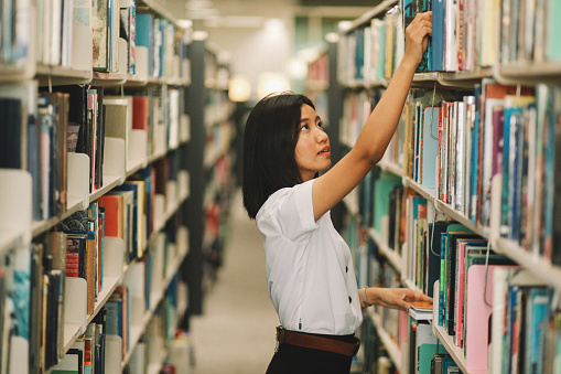 Young Asian student searching through the shelves of the library, searching for books for her studies.