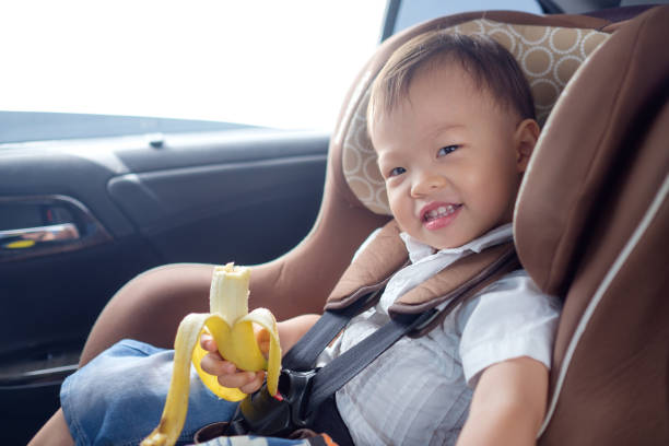 cute little asian 18 months / 1 year old toddler baby boy child sitting in safety carseat holding & enjoy eating banana - car baby baby car seat child imagens e fotografias de stock